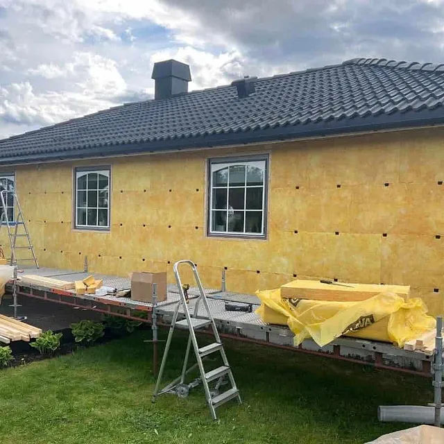 Bolig med gul isolasjon montert på yttervegg, stillas og materialer foran huset.