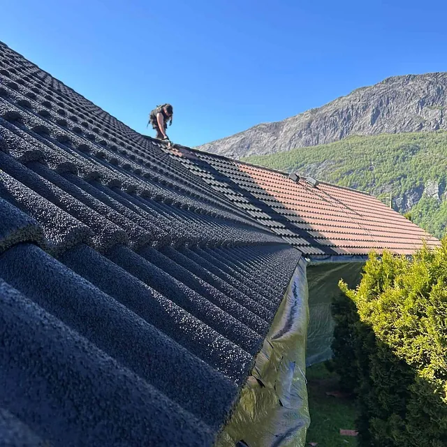 Arbeider på skrått hustak legger ny mørk takstein med fjellandskap i bakgrunnen.