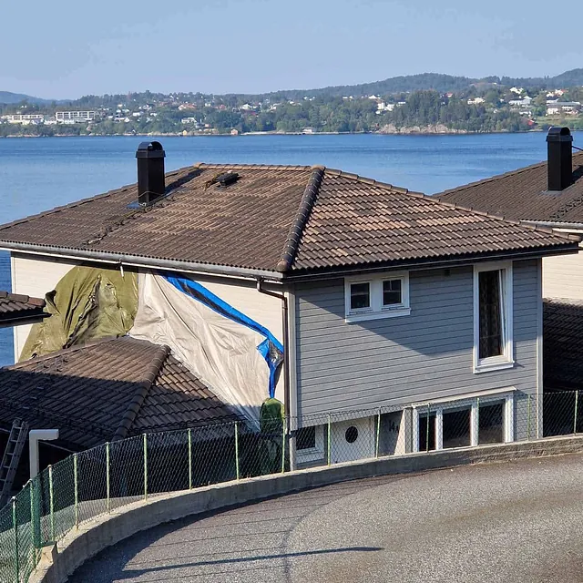 Hus ved sjøen med deler av tak og vegg dekket av presenning under arbeid.
