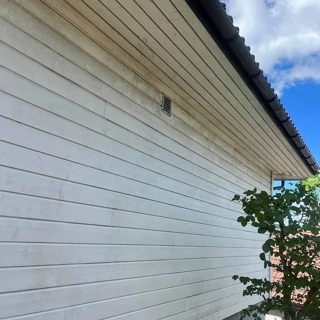 Hvit husvegg med skitt og misfarging under takutstikket, sett fra bakkenivå.